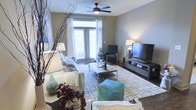 This single-floor space centers around a thoughtfully designed living room that balances comfort and style. Large double doors with sheer white curtains and blinds allow an abundance of natural light to flood the room, creating a warm and inviting atmosphere. The living area is anchored by a plush sofa adorned with decorative pillows and a cozy throw blanket, complemented by an accent armchair and a matching seated cube in teal. A sleek rectangular coffee table sits atop a patterned area rug, tying the seating arrangement together. The entertainment setup features a large flat-screen television mounted on a modern low-profile media console, providing a functional focal point for the room. Overhead, a classic ceiling fan with a built-in light fixture provides both airflow and ambient lighting. Decorative touches throughout — including sculptural branches, table lamps, and floral accents — add character and depth to the space. The hard flooring, visible along the perimeter, contrasts nicely with the soft area rug, contributing to the overall polished aesthetic. The space is well-suited for both relaxed everyday living and casual entertaining.