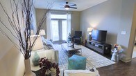 This single-floor space centers around a thoughtfully designed living room that balances comfort and style. Large double doors with sheer white curtains and blinds allow an abundance of natural light to flood the room, creating a warm and inviting atmosphere. The living area is anchored by a plush sofa adorned with decorative pillows and a cozy throw blanket, complemented by an accent armchair and a matching seated cube in teal. A sleek rectangular coffee table sits atop a patterned area rug, tying the seating arrangement together. The entertainment setup features a large flat-screen television mounted on a modern low-profile media console, providing a functional focal point for the room. Overhead, a classic ceiling fan with a built-in light fixture provides both airflow and ambient lighting. Decorative touches throughout — including sculptural branches, table lamps, and floral accents — add character and depth to the space. The hard flooring, visible along the perimeter, contrasts nicely with the soft area rug, contributing to the overall polished aesthetic. The space is well-suited for both relaxed everyday living and casual entertaining.