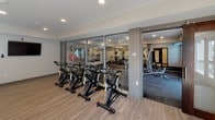 This single-floor fitness studio is thoughtfully designed to accommodate a variety of workout styles in a bright, open environment. The space is finished with light hardwood-style flooring throughout, contributing to a clean and modern aesthetic that is both functional and visually welcoming.

The spinning area is outfitted with a row of stationary cycling bikes and is positioned adjacent to a large glass partition wall that connects to a separate weight and cardio room. This adjoining space is visible through the glass, housing treadmills, an elliptical, and strength training equipment, creating a seamlessly integrated fitness environment.

A dedicated open exercise zone occupies a generous portion of the studio, offering ample room for stretching, yoga, or group fitness activities. A wall-mounted yoga mat rack keeps equipment neatly organized. One wall features a deep teal accent panel anchoring a full-length ballet barre and large floor-to-ceiling mirror, ideal for dance or barre-style workouts.

A wall-mounted flat-screen TV provides entertainment or instructional support. Recessed lighting and multiple exterior glass doors flood the space with natural light, while direct access to an outdoor area further enhances the open, airy atmosphere of this well-equipped facility.