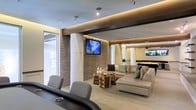 This single-floor space showcases a sophisticated, open-concept design that blends modern materials with thoughtful lighting. A striking textured stone accent wall serves as the focal point of the lounge area, anchoring a wall-mounted flat-screen television and providing rich visual contrast against the smooth, neutral-toned surfaces throughout. Structural columns with a curved, sculpted finish add architectural interest while subtly defining different zones within the open layout.

The lounge area is furnished with a generously sized sectional sofa in a warm beige tone, complemented by teal accent pillows and a distinctive rustic wood coffee table that introduces an organic element amid the contemporary design. Recessed track lighting and elegant cove lighting embedded in the ceiling create a layered, ambient atmosphere, casting a warm golden glow across the space.

Adjacent areas are partially visible, hinting at additional functional zones, including what appears to be a bar or counter area with pendant lighting. Large windows allow natural light to filter in, enhancing the open, airy feel. The overall ambiance is polished and inviting, suitable for both relaxation and social gatherings.