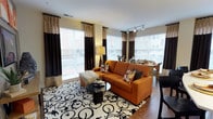 This single-floor space showcases a thoughtfully designed open-concept layout that seamlessly connects the living and dining areas, creating a welcoming and functional environment. The living area is anchored by a bold burnt-orange sofa paired with a striking black-and-white patterned area rug, lending a vibrant, contemporary aesthetic to the room. A sleek glass coffee table and coordinating accent chair complete the seating arrangement, offering both style and comfort.

Large windows dressed with elegant two-tone curtains — featuring rich chocolate brown and warm neutral panels — flood the space with abundant natural light while framing views of the surroundings. The windows are fitted with horizontal blinds, allowing for versatile light control throughout the day. Track lighting on the ceiling provides additional illumination, enhancing the bright and airy ambiance.

The dining area transitions naturally from the living space, featuring a wooden dining table and chairs that complement the warm hardwood-style flooring running throughout. The open flow between zones makes the space feel expansive and well-proportioned. The neutral wall tones serve as a clean backdrop that allows the colorful furnishings and decorative accents to stand out, resulting in a space that is both visually dynamic and inviting.