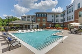 Located on Atlanta Student Movement Boulevard SW in Atlanta, Georgia, this single-floor residential unit is part of a well-appointed multi-story community with classic brick and contemporary exterior architecture. The interior features a clean, neutral color palette with crisp white walls and white panel doors throughout. Living areas are finished with plush wall-to-wall carpeting that provides comfort underfoot, while transitional spaces showcase rich, dark hardwood-style flooring for a refined contrast. Windows fitted with horizontal blinds allow for adjustable natural light across the rooms, contributing to a bright and airy atmosphere. The layout is open and functional, with well-proportioned rooms offering flexibility for a variety of furnishing arrangements. The community's outdoor amenity area is a standout feature, centered around a large, sparkling swimming pool with crystal-clear water. A row of white lounge chairs lines the pool deck, ideal for relaxing in the sun. A covered wooden pergola cabana with seating provides a shaded gathering space, all enclosed within an ornamental iron fence. Lush landscaping and decorative lighting complete the outdoor environment, creating an inviting retreat. The property's location in Atlanta's historic west side neighborhood adds further context to this well-maintained residential community.