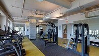 Located at 202 N West St in Raleigh, NC, this fitness center offers a well-equipped, single-floor workout facility designed to accommodate a wide range of exercise needs. The space features an open layout with an industrial aesthetic, highlighted by exposed ductwork and structural columns painted white, lending the room a modern, utilitarian character. Overhead fluorescent lighting ensures the entire floor is evenly and brightly illuminated throughout.

The gym floor is covered in durable rubber flooring with a distinctive yellow and black speckled pattern, providing both safety and comfort during workouts. A comprehensive selection of strength-training machines is arranged throughout the space, including cable machines, multi-functional weight stations, and a variety of resistance equipment suited for both upper and lower body training. Large mirrors line one wall, enhancing the sense of space while allowing users to monitor their form during exercise.

Ceiling-mounted fans contribute to air circulation across the facility, supporting a comfortable environment during intense workouts. Natural light filters in through windows along the perimeter, adding to the bright and energizing atmosphere. An exit door with signage provides clear safety access. Overall, this fitness center is a functional, well-maintained space built to support a active and health-focused lifestyle.