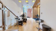 Located at 625 Avenue de l'Equinoxe in Brossard, Quebec, this contemporary two-floor fitness facility is thoughtfully designed to support a wide range of exercise activities. The main floor houses a spacious exercise room finished with warm hardwood flooring and accented by a striking wood-paneled feature wall with integrated lighting. Distinctive yellow pendant lights hang from the ceiling, adding a vibrant, modern touch to the space. Large floor-to-ceiling windows flood the area with natural light while offering views of the surrounding urban landscape. The room is equipped with cardio machines including treadmills, ellipticals, stationary bikes, and a rowing machine, as well as a multi-function cable machine, a weight bench, and a comprehensive dumbbell rack. Full-length mirrors extend the sense of space. A staircase with glass railings connects to the upper level, which features a dedicated studio or multipurpose room with light wood-toned flooring, recessed lighting, and frosted sliding glass panel partitions that allow soft, diffused light to pass through. This airy, open room is well-suited for yoga, stretching, or group fitness, and is stocked with exercise mats, stability balls, and other accessories. A hallway and additional unclassified rooms complete the layout, supporting smooth circulation and functionality throughout the facility.