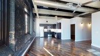 This loft at Bradford Mills Lofts showcases a thoughtful blend of industrial character and modern design. The open-concept kitchen serves as a central feature, outfitted with dark cabinetry, stainless steel appliances including a range, microwave, dishwasher, and refrigerator, and a white subway tile backsplash that provides a clean contrast to the darker finishes. A double-basin sink anchors the peninsula, adding functionality to the layout.

Overhead, the original wood-plank ceiling with exposed structural beams and visible ductwork reinforces the building's industrial loft heritage. Pendant lighting and a ceiling fan complement the vertical volume created by the soaring ceiling height. Original grid markings on the structural columns — a nod to the building's mill history — add authentic character to the space.

Underfoot, richly toned, wide-plank hardwood floors run throughout, adding warmth against the otherwise raw industrial backdrop. A hallway leading from the main living area hints at additional rooms beyond, while white walls keep the space feeling bright and open. The overall ambiance balances historic mill aesthetics with contemporary residential comfort.