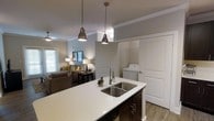 This single-floor residence offers a thoughtfully designed open-concept layout that seamlessly connects the kitchen and living areas. The kitchen serves as a central hub, anchored by a spacious island with a crisp white quartz countertop and a deep double-basin stainless steel sink. Dark espresso cabinetry beneath the island provides a striking contrast to the light surfaces above. Two polished metal pendant lights hang overhead, adding an industrial-chic touch that complements the overall aesthetic. Crown molding runs throughout, lending an air of refinement to the space. Adjacent to the kitchen, an in-unit washer and dryer area is neatly tucked behind panel-style doors, offering convenient, discreet laundry access. The living area flows naturally from the kitchen, featuring warm, layered lighting from table lamps and a ceiling fan, alongside a comfortable seating arrangement. Natural light filters in through a window dressed with horizontal blinds, brightening the neutral gray walls and light wood-look flooring. The cohesive color palette of soft grays, whites, and warm accents creates an inviting and functional atmosphere throughout this well-finished single-level home.