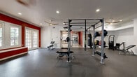 This expansive fitness center offers a comprehensively outfitted workout facility spread across a single floor, encompassing multiple distinct training zones designed to support a wide range of exercise disciplines. The main cardio area features a full row of modern treadmills, ellipticals, and other cardio machines, complemented by floor-to-ceiling mirrors that enhance the sense of space and allow for form monitoring. Red accent walls add a bold, energizing visual identity throughout the space, contrasting sharply with the clean white walls and dark rubber flooring underfoot. A dedicated strength and free weights zone includes a multi-station power rack, adjustable benches, a complete dumbbell rack, and a cable machine, with decorative mandala wall art providing an unexpected design touch. A separate functional training room houses a large freestanding rig with pull-up bars, a heavy punching bag, suspension trainers, medicine balls, and kettlebells. Ceiling fans and recessed lighting ensure comfort throughout. A secondary area offers cardio equipment alongside a water fountain and informational displays. The facility also connects to a bright lounge with large windows overlooking a courtyard, and an outdoor colonnade features striking natural stone arched columns with terracotta tile flooring, providing a welcoming entrance to the complex.