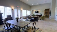 This single-story space offers an open-concept layout that seamlessly blends a dining area with a comfortable lounge, creating a versatile and inviting environment. The dining area features a sturdy wood farmhouse-style table paired with industrial-accented chairs, anchored by a richly patterned area rug that adds visual interest to the light tile flooring beneath.

The high ceilings amplify the sense of spaciousness throughout, while recessed lighting and wall-mounted sconces provide warm, layered illumination. Large windows along one wall allow abundant natural light to filter in, enhancing the room's airy feel. A set of white French doors offers easy access to the exterior.

A graceful arched opening defines the transition into the lounge area, where dark leather sofas and green upholstered accent chairs are arranged around a wall-mounted flat-screen television. A dark wood console table serves as a decorative focal point, styled with lamps, greenery, and decorative accents. The neutral warm-gray wall tones and carefully chosen furnishings throughout contribute to a cohesive, sophisticated ambiance that balances functionality with comfort.
