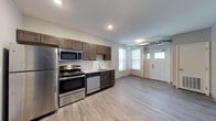 This well-appointed single-floor space offers a clean, modern aesthetic defined by an open-concept layout that seamlessly connects the kitchen and living areas. The kitchen is a standout feature, equipped with a full suite of stainless steel appliances including a refrigerator, electric range, microwave, and dishwasher. Rich, dark wood cabinetry provides ample storage and pairs stylishly with light countertops and a classic white tile backsplash. A stainless steel sink with a modern faucet sits beneath a bright window, allowing natural light to filter into the space and offer views of the surrounding neighborhood. Light-toned hardwood-style flooring extends throughout, lending warmth and continuity to the open floor plan. Recessed lighting overhead ensures the space is well-lit throughout the day and evening. The generous open area adjacent to the kitchen provides flexible space for dining or lounging. A private entrance with a white paneled door adds convenience and character. A hallway visible from the main living area leads to a bathroom featuring matching dark wood cabinetry, maintaining a cohesive design throughout. The overall ambiance is bright, clean, and move-in ready.