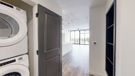 This thoughtfully designed 2-bedroom, 2-bathroom unit showcases an industrial-modern aesthetic that is immediately evident upon entry. Light wood-tone flooring flows throughout the open layout, complemented by exposed concrete ceilings that lend an urban, loft-like character to the space. Charcoal-gray two-panel doors add a refined contrast against the crisp white walls.

The living area is a standout, featuring dramatic floor-to-ceiling windows with black frames that flood the room with natural light and open to a private balcony with city views. A ceiling fan adds functionality to this bright, airy space. The kitchen is well-appointed with dark cabinetry, white quartz countertops, stainless steel appliances including a refrigerator, range, and microwave, and a central island that provides additional prep and seating space. Pendant lighting adds a modern finishing touch.

The primary bedroom offers generous proportions with access to an en-suite bathroom and a spacious walk-in closet featuring wire shelving. The bathrooms are finished in a contemporary style, with dark vanity cabinetry, gold hardware accents, white quartz countertops, and large framed mirrors. Both bedrooms benefit from the same consistent flooring and elevated ceiling treatment, maintaining a cohesive design throughout.