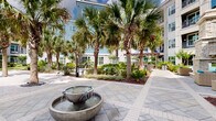 The outdoor courtyard at 1400 Encore Pl in Sanford, Florida, is a thoughtfully designed communal space that serves as the central gathering area of a modern residential community. A decorative bowl fountain anchors the main entry plaza, set atop a patterned paver courtyard that creates an inviting first impression. Stone-accented columns and warm wood-toned entry gates frame the building's ground-floor entrance, lending an upscale aesthetic to the arrival experience.

Winding paver pathways connect the various zones of the courtyard, guiding residents through dense tropical landscaping filled with palms, ornamental shrubs, and vibrant ground cover. The lush plantings provide a sense of privacy and a distinctly Florida-resort atmosphere throughout the space.

A pergola-covered seating area offers a shaded spot for outdoor dining and relaxation, furnished with tables and chairs beneath a rich wood-toned overhead structure. Nearby, a wicker lounge seating area with teal cushions provides a casual retreat surrounded by verdant garden beds. One of the standout amenities is a putting green, offering a recreational option within the courtyard's open sandy section. Swing chairs and benches are scattered throughout, enhancing the relaxed, resort-like ambiance. The multi-story building wraps around the courtyard, with balconies overlooking the well-maintained grounds below.