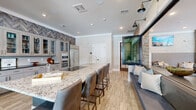 This well-appointed kitchen is a masterclass in contemporary design, offering a thoughtfully curated blend of textures, materials, and functional elegance. At its center stands a striking large island topped with white and gray granite, accented by a bold teal-painted base and surrounded by leather upholstered bar seating — ideal for casual dining or entertaining.

The perimeter cabinetry features floor-to-ceiling glass-front upper cabinets in a soft gray finish, paired with a white subway tile backsplash and dark countertops. A dramatic herringbone wood-panel accent wall runs the full length of the cabinet wall, adding warmth and visual depth. Stainless steel appliances, including a full-size refrigerator, double wall oven, and microwave, provide a sleek, professional-grade cooking environment. Recessed lighting and industrial-style wall sconces illuminate the space with both task and ambient light.

Wood-look tile flooring runs throughout, offering a warm, natural aesthetic underfoot. A standout architectural feature is the large folding glass and metal pass-through window that opens the kitchen directly to an adjacent lounge area, framed by stacked stone columns. A built-in upholstered bench with decorative pillows sits beside it, seamlessly connecting the two spaces. Crown molding and clean white walls complete the polished, modern-transitional aesthetic.