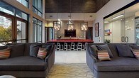 This thoughtfully designed single-floor amenity space in Nashville's vibrant urban core offers a range of distinct areas tailored for both productivity and leisure. The centerpiece lounge features soaring double-height ceilings with floor-to-ceiling windows that flood the space with natural light, complemented by a striking red-topped bar illuminated by a trio of industrial-style cage pendant lights. Plush gray sofas and a patterned area rug anchor the seating area, while a built-in fireplace set within a large tile surround provides warmth alongside a wall-mounted television. A dedicated conference room showcases a live-edge wood table, sculptural chandelier, and bold artwork, creating an inspiring setting for meetings. The game room features a pool table, vibrant orange-accented carpet, booth-style seating, and direct access to an outdoor area. A compact yet functional workstation nook with patterned wallcovering and modern furnishings serves as a private business center. Throughout, the design palette combines warm wood tones, textured wall treatments, and bold orange accents, resulting in a cohesive, contemporary atmosphere with an energetic yet sophisticated character.