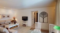 This single-floor space presents an open, cohesive layout that seamlessly connects the living and dining areas. The living area is finished with neutral-toned carpet flooring and smooth white walls, creating a clean and understated backdrop. A white media console holds a flat-screen television and features built-in drawers and lower shelving with woven baskets, offering both style and storage. A white coffee table with matching wicker basket storage anchors the seating area, complementing the overall light color palette.

Adjacent to the living space, a glass-top dining table is paired with white slatted dining chairs, adding a casual yet refined touch. A striking decorative metal tree wall sculpture serves as a prominent focal point on the wall, adding an artistic, nature-inspired element to the room. A classic table lamp provides warm ambient lighting to complement the natural light in the space.

A louvered closet door and an open doorway leading to an adjoining hallway or room are visible, suggesting practical storage options and connectivity to additional areas. The overall ambiance is light, airy, and welcoming, characterized by consistent white tones, warm textures, and tasteful decorative accents.