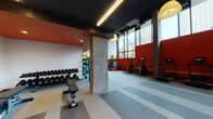 Located at 1150 Clay St in Oakland, California, this contemporary fitness center is designed to support a full range of workout routines in a well-equipped, single-floor facility. The space makes a strong visual impression with bold red accent walls contrasting against a dark ceiling, exposed concrete columns, and striped rubber flooring that defines distinct workout zones throughout the gym.

Natural light pours in through expansive floor-to-ceiling windows along one wall, offering views of the surrounding Oakland cityscape and creating an open, energizing atmosphere. The high ceilings amplify the sense of space, while industrial-style linear lighting and pendant fixtures supplement the natural light throughout the interior.

The cardio zone features multiple treadmills and elliptical machines positioned along the windowed wall, allowing users to exercise with city views. Adjacent to this area, a well-stocked free weights section includes a full rack of dumbbells, stability balls in various sizes, plyo boxes, and resistance training accessories. A wall-mounted mirror expands the visual depth of this section, while a flat-screen TV is mounted overhead for entertainment.

The strength training area houses a cable machine system and a squat rack with barbell equipment, catering to a variety of resistance training needs. A stainless steel water station is conveniently located near the entrance, which features sleek glass double doors. The overall design balances functionality with an energetic, modern aesthetic.
