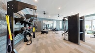 This fitness center offers a thoughtfully designed, single-floor layout encompassing a generous amount of workout space. The interior is unified by a distinctive blue marbled wallcovering that adds a cohesive, modern aesthetic throughout. Patterned carpet tiles cover the main fitness floor, providing durability and visual interest, while recessed lighting and ceiling fans maintain a comfortable atmosphere.

The main workout area is well-equipped with True-brand strength machines, TRX suspension training rigs stocked with medicine balls and battle ropes, adjustable benches, and a full dumbbell rack. Large wall-mounted mirrors amplify the sense of space and allow users to monitor their form. A dedicated cardio zone features treadmills, ellipticals, and stair climbers positioned near oversized windows that invite natural light and outdoor views.

A separate yoga room offers a distinct environment with hardwood flooring, a richly colored tray ceiling, floor-to-ceiling mirrors, and multiple windows. Spin bikes and balance trainers are available within this space for diverse mind-body workouts.

Near the entrance, a built-in storage unit with open cubbies and wicker baskets provides organized storage for yoga mats and accessories, complemented by an elevator for convenient access.