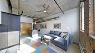 This single-floor space embraces an industrial-modern aesthetic, characterized by its striking exposed concrete ceiling with visible ductwork and conduit piping — hallmark features of loft-style design. A ceiling fan is centrally mounted, adding both function and visual continuity to the raw overhead finish.

The open-plan layout seamlessly connects the living area to the kitchen zone. The living space is anchored by a sectional sofa in slate grey, positioned around a low-profile coffee table and accented by a vibrant multicolored area rug that introduces warmth and contrast against the polished concrete flooring. A floor lamp and a curated gallery wall of framed artwork add personality and a lived-in character to the space.

The kitchen area, partially visible, features a dark-toned island or peninsula with bar stool seating and pendant lighting suspended above. Stainless steel appliances and warm wood cabinetry provide a balanced contrast to the otherwise cool industrial palette. Natural light enters from one side, brightening the concrete floors and enhancing the open, airy feel. Overall, the space conveys a cohesive urban loft sensibility with a comfortable, contemporary edge.