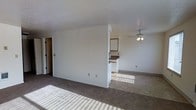 This single-floor space features an open, functional layout designed to maximize usable living area. The main living space is finished with wall-to-wall carpet, providing a comfortable and warm feel underfoot. A ceiling fan is centrally positioned overhead, offering both air circulation and a modest lighting fixture for the room.

A partial dividing wall separates the living area from the kitchen zone, creating a sense of distinction between spaces while maintaining an open feel. The kitchen area transitions to tile flooring, a practical choice for a food-preparation zone. White upper cabinetry is visible beyond the dividing wall, along with what appears to be a countertop with a darker surface and standard white appliances, providing essential functionality.

The textured ceiling adds a classic touch throughout the space. Neutral white walls keep the interior feeling bright and clean, offering a blank canvas for personalization. Natural light filters into the space, illuminating both the carpeted and tiled flooring areas. A doorway is visible along one wall, indicating access to additional rooms or storage. Overall, the space presents a straightforward, practical layout suited for everyday living.