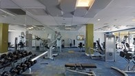 This fitness center offers a spacious, single-level layout designed to accommodate a comprehensive workout experience. The room is generously proportioned, providing ample space between equipment stations to ensure comfortable movement throughout the facility.

The strength training area is well-stocked with a variety of cable and resistance machines positioned throughout the space, allowing users to target multiple muscle groups. Free weights, including a dumbbell rack, are also available along one side of the room. Cardio equipment, including rowing machines and other aerobic stations, is visible toward the perimeter of the space.

Floor-to-ceiling mirrors line one wall, enhancing the sense of openness while allowing users to monitor their form during exercise. A flat-screen television is mounted on the wall, providing entertainment during workouts.

The ceiling features a distinctive coffered tile design with integrated recessed lighting, contributing to a bright and modern ambiance. A bold yellow-green accent column adds a pop of color to the otherwise neutral interior palette. The floor is finished with a patterned, multi-tone carpet that adds visual texture while providing underfoot comfort throughout the facility.