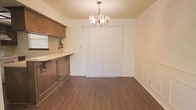 This single-floor residence offers a straightforward and functional layout with a warm, inviting ambiance throughout. The dining area serves as a central gathering space, highlighted by rich, dark hardwood-style flooring that runs seamlessly across the room. A classic chandelier hangs overhead, providing elegant accent lighting and lending a touch of traditional charm to the space. Vertical blinds covering a large sliding door or window allow for adjustable natural light and privacy, while neutral cream-toned walls keep the atmosphere bright and open. Adjacent to the dining area, the kitchen features dark wood cabinetry with a clean countertop surface, offering practical storage and prep space. A range hood is mounted above the cooking area, and a fire extinguisher is conveniently accessible, reflecting thoughtful safety considerations. The kitchen's layout allows for easy interaction with the dining space, creating a connected and efficient flow. Subtle wainscoting detail along the lower portion of the dining room wall adds a classic architectural touch. Overall, this residence combines practical functionality with understated traditional design elements for comfortable everyday living.