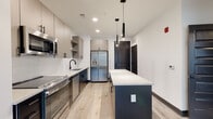 This thoughtfully designed single-floor unit showcases a cohesive modern aesthetic with high-quality finishes throughout. The open-concept kitchen serves as the centerpiece, featuring two-tone cabinetry with warm taupe uppers and dark lower cabinets, a white subway tile backsplash, and a full suite of stainless steel appliances including a side-by-side refrigerator with ice and water dispenser, electric range, dishwasher, and built-in microwave. A large kitchen island with a white quartz countertop provides additional workspace and seating, while matte black pendant lights add a striking design accent overhead. Light wide-plank hardwood flooring flows seamlessly from the kitchen into the open living areas, which are bathed in natural light from oversized windows with outdoor views. The bedrooms continue the clean, bright aesthetic with the same hardwood flooring, dark baseboards, and generous window arrangements. The bathroom is finished with large-format tile flooring, a wide wood-toned vanity with quartz countertop, a framed mirror, and matte black fixtures. Recessed and track lighting throughout ensures well-illuminated spaces. The overall ambiance is clean, contemporary, and move-in ready.