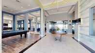 This generously sized single-floor amenity space offers a well-appointed layout designed for both socializing and relaxation. The main entry area features large-format white tile flooring, structural columns with blue and gray accents, and a striking geometric gold chandelier overhead. A central round wood-top table with a chrome base anchors the open foyer, while frosted glass partitions add a layer of privacy and architectural interest.

The adjoining game and lounge area features warm hardwood-style flooring, a full-size billiard table, and comfortable seating arrangements with velvet upholstered chairs in rich jewel tones. The living room area is highlighted by soaring vaulted ceilings, a wall-mounted fireplace, a large flat-screen TV, and French doors that invite abundant natural light and outdoor views. A bold teal herringbone accent wall and a circular pendant light fixture lend the space a sophisticated, contemporary character.

The dining bar area accommodates multiple guests at a sleek counter with upholstered bar-height chairs in gold-toned metal frames, flanked by tall windows that flood the space with natural light. Deep blue textured wallcovering and curated wall art complete the refined aesthetic. A wide, well-lit hallway with wall sconces connects the various zones, ensuring smooth flow throughout the entire space.
