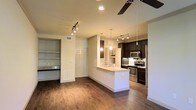 Located in North Richland Hills, Texas, this single-floor residence offers a well-designed open-concept layout that seamlessly connects the living and kitchen areas. Rich, dark hardwood-style flooring runs throughout the main living space, adding warmth and a sense of continuity to the interior.

The kitchen is a standout feature, outfitted with dark espresso cabinetry, stainless steel appliances including a range, microwave, and refrigerator, and a peninsula with a light-toned countertop that doubles as a breakfast bar. Pendant lighting hangs above the peninsula, complementing the track lighting that illuminates the space with a modern, layered lighting scheme. A ceiling fan overhead adds both function and style to the open living area.

Adjacent to the kitchen, a built-in shelving niche provides practical display or storage space, while a nearby door leads to additional areas of the home. Recessed lighting in the ceiling enhances the bright, clean aesthetic throughout. The neutral wall tones paired with the contrasting dark cabinetry and flooring create a balanced, contemporary ambiance. Overall, the space offers a functional and stylish environment well-suited for everyday living.