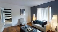 This single-floor residence offers a thoughtfully organized layout that balances comfort and functionality. The living area serves as the centerpiece of the space, showcasing rich, warm hardwood flooring that extends throughout the main areas. A bold dark accent wall adds depth and character to the room, complementing the neutral white walls and creating a modern, stylish ambiance. A large window dressed with vertical blinds allows natural light to filter in, brightening the space and adding an airy feel. The living area is well-suited for relaxed gatherings, with ample room for seating and a coffee table centerpiece.

Adjacent to the living area, the kitchen is efficiently laid out with tile flooring, providing a clean and practical workspace equipped with essential appliances. The transition between the kitchen and living spaces is seamless, lending an open, connected feel to the overall floor plan.

A dedicated sleeping area is situated toward the rear of the unit, offering a degree of separation and privacy from the main living space. A ceiling fan overhead adds both comfort and functionality to this area.

The space also includes a bathroom and entryway corridor, completing a compact yet well-structured layout that makes excellent use of every square foot.