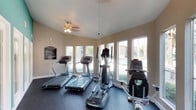 This single-story space presents two distinct areas that together offer both functional and aesthetic appeal. The fitness center is well-equipped with multiple treadmills, an elliptical trainer, and a stationary bike, all arranged on protective rubber flooring. A ceiling fan provides ventilation, while a row of large windows along one wall floods the room with natural light and offers views of an outdoor pool area, creating an inviting environment for exercise. Fitness center rules are posted near the entrance for resident reference.

The dining area demonstrates a carefully curated interior design sensibility. A striking round green marble pedestal table serves as the focal point, surrounded by woven rattan chairs and anchored by a textured area rug over dark concrete flooring. A large-framed abstract artwork adorns the neutral wall, complementing a sculptural grey urn centerpiece on the table. A sliding barn door connects to an adjacent space, adding an industrial touch. Recessed lighting provides a clean, modern overhead illumination. The overall ambiance blends artistic character with understated elegance throughout.