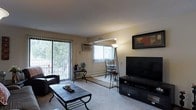 This single-floor space showcases a thoughtfully arranged living room as its central feature. The room is finished with neutral beige walls and a textured ceiling, creating a warm and inviting atmosphere. Soft, wall-to-wall carpeting covers most of the floor, complemented by a tiled-surface coffee table with a decorative metal frame that adds a rustic touch.

Seating includes a dark brown sofa adorned with a patterned accent pillow and a matching leather recliner draped with a cozy throw blanket, both positioned to face the entertainment area. A large flat-screen television sits atop a dark media console along one wall, accompanied by a framed art piece above it.

The living room benefits from generous natural light, primarily through a large sliding glass door fitted with vertical blinds, which opens to an outdoor balcony area surrounded by greenery. An additional window further brightens the space. A wall-mounted air conditioning unit provides climate control. Decorative metallic wall art in rich amber and red tones adds personality and visual interest throughout the room, tying the warm color palette together cohesively.