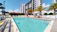 This outdoor amenity space offers a resort-style experience centered around a generously sized swimming pool with pristine, turquoise water. The pool area is thoughtfully designed with a wide, clean sundeck that provides ample room for lounging and relaxation. Comfortable chaise lounge chairs with bright yellow cushions are positioned along the poolside, inviting residents and guests to unwind in the sun. Tall palm trees add a tropical ambiance, offering natural shade and a lush, coastal aesthetic that complements the open-air setting. The surrounding environment is bright and airy, benefiting from abundant natural sunlight and open sky views. A sleek utility structure is situated at the far end of the pool deck, blending seamlessly into the overall design. In the background, a striking multi-story residential tower rises prominently, featuring a modern façade with clean lines, balconies, and a predominantly white and dark-accented exterior. Flowering shrubs and greenery accent the perimeter, adding color and vibrancy to the space. The overall atmosphere is lively yet relaxing, making this pool area an ideal outdoor retreat with a distinctly warm, Florida-inspired character.