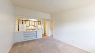This single-floor unit offers a functional and straightforward layout connecting several well-defined areas. The living room is generously proportioned, finished with neutral-toned carpet and crisp white walls beneath a textured ceiling. It opens toward the kitchen via a distinctive pass-through divider equipped with built-in shelving, creating a semi-open connection between the two spaces without fully removing the separation.

The kitchen features wood-trimmed white cabinetry extending to the upper walls, a stainless steel sink, light laminate countertops, and hardwood-style flooring. White appliances, including a refrigerator and dishwasher, are integrated throughout. An overhead skylight provides additional brightness to the kitchen area.

The bedrooms are carpeted and well-lit, with one room featuring a ceiling fan and a large window fitted with vertical blinds that allows natural outdoor views and ample daylight. A second carpeted bedroom offers a quiet, neutral environment with a floor vent for climate control.

The full bathroom includes a single-sink vanity with cabinet storage, a combined tub and shower, and hardwood-style flooring. A dedicated laundry room accommodates a full-sized washer and dryer side by side, with a water heater also housed within the space.