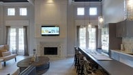 This single-story home showcases an open-concept layout that seamlessly connects the living and dining areas into one cohesive, elegant space. The living area is anchored by a striking fireplace clad in a textured tile surround, framed by crisp white wainscoting and paneling that extends dramatically to the ceiling, creating a sophisticated focal point. A wall-mounted television sits neatly above the mantel, blending modern convenience with classic design. The soaring ceiling height amplifies the sense of grandeur, drawing the eye upward and allowing abundant natural light to pour in through a set of tall upper windows. Adjacent to the living area, a substantial dining table with richly upholstered high-back chairs provides generous seating. Above it, a trio of glass pendant lights with exposed bulbs adds a contemporary industrial touch that contrasts beautifully with the traditional architectural details. Large windows along the dining area frame views of the outdoors, further brightening the interior. Light hardwood flooring runs throughout, complementing the neutral gray walls and white trim. The overall ambiance is one of refined comfort—elegant yet approachable, with thoughtful design details throughout.