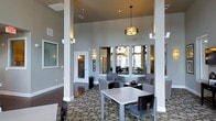This well-appointed single-floor communal space exudes a modern, welcoming ambiance designed for comfort and functionality. The interior is defined by impressively high ceilings that create an open, airy atmosphere, anchored by bold structural columns finished in crisp white that provide architectural character throughout the room. A statement drum pendant light hangs centrally, complemented by wall sconces that cast a warm, inviting glow across the space.

The flooring features a richly patterned carpet with a bold circular motif, adding visual interest and a sense of sophistication. Seating is arranged thoughtfully throughout, with modern dining-height tables paired with sleek, contemporary chairs offering casual gathering spots, while upholstered lounge seating along the walls provides a more relaxed setting.

The walls are painted in a warm taupe tone, lending a neutral, refined backdrop. Large windows allow natural light to filter into adjacent areas visible through interior glass partitions, connecting the main lounge to additional rooms beyond. Frosted glass door panels provide privacy while maintaining a sense of openness. The overall design balances elegance with practicality, making this a versatile and inviting space well-suited for social gatherings and everyday use.