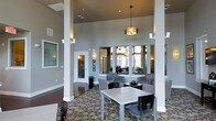 This well-appointed single-floor communal space exudes a modern, welcoming ambiance designed for comfort and functionality. The interior is defined by impressively high ceilings that create an open, airy atmosphere, anchored by bold structural columns finished in crisp white that provide architectural character throughout the room. A statement drum pendant light hangs centrally, complemented by wall sconces that cast a warm, inviting glow across the space.

The flooring features a richly patterned carpet with a bold circular motif, adding visual interest and a sense of sophistication. Seating is arranged thoughtfully throughout, with modern dining-height tables paired with sleek, contemporary chairs offering casual gathering spots, while upholstered lounge seating along the walls provides a more relaxed setting.

The walls are painted in a warm taupe tone, lending a neutral, refined backdrop. Large windows allow natural light to filter into adjacent areas visible through interior glass partitions, connecting the main lounge to additional rooms beyond. Frosted glass door panels provide privacy while maintaining a sense of openness. The overall design balances elegance with practicality, making this a versatile and inviting space well-suited for social gatherings and everyday use.