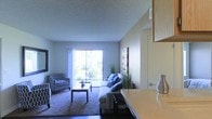 This single-floor residence features an open-concept layout that connects the kitchen and living areas, creating a cohesive and functional living environment. The living room is carpeted and comfortably furnished with a sofa and a patterned accent chair arranged around a central coffee table. A large sliding glass door floods the space with natural light and provides views of the greenery outside, enhancing the room's airy atmosphere. Decorative touches, including a tall potted plant and wall art, add warmth to the neutral-toned walls beneath a textured ceiling. Adjacent to the living area, the kitchen features light wood cabinetry and a countertop that extends toward the living space, offering a natural transition between the two areas. The overall ambiance is clean, functional, and welcoming, with an emphasis on natural light and a practical open-plan design that maximizes the sense of space throughout the interior.