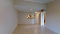 This single-floor residence offers a clean, functional layout designed to maximize comfort and usability. The main living area is spacious and open, finished with light-toned ceramic tile flooring that extends throughout the space, lending a cohesive and airy feel. Crown molding along the ceiling adds a subtle touch of elegance, while recessed lighting provides even, practical illumination.

The kitchen is accessible via a convenient pass-through window and counter opening, connecting it visually and functionally to the living area. It features warm wood-toned cabinetry, a stainless steel range, a sink positioned beneath the pass-through, and tile backsplash, making it a compact yet well-equipped cooking space.

A hallway leads from the main living area to additional rooms, including what appears to be a bathroom and further living spaces, all finished in consistent neutral tones. The open doorway visible from the living area reveals a similarly tiled secondary space, hinting at a well-connected floor plan.

Overall, the space presents a bright, neutral palette with warm accents, practical finishes, and a straightforward layout that is both easy to navigate and comfortable for everyday living.