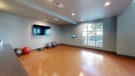 Located at 1700 Newbury Park Dr in San Jose, California, this two-floor fitness center offers a comprehensive workout facility designed with both function and aesthetics in mind. The main floor serves as the primary exercise area, characterized by gleaming hardwood floors, soaring ceilings, and large multi-pane windows that flood the space with natural light and offer views of the surrounding greenery. Structural columns punctuate the open floor plan, adding an architectural dimension to the layout. The main workout area is well-appointed with cable machines, treadmills, ellipticals, free weights, and weight benches, all reflected in large wall-mounted mirrors that enhance the sense of openness. A dedicated studio area on the main floor features hardwood flooring, a wall-mounted flat-screen TV, and stability balls, ideal for stretching, yoga, or group exercise. A staircase leads to the mezzanine level, which offers a carpeted secondary workout zone equipped with cardio machines and mirror accents. Colorful wall art adds a modern, energetic touch throughout. Recessed lighting and pendant fixtures provide warm, even illumination, while the overall neutral color palette of soft grays and warm wood tones creates a clean, motivating environment.