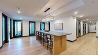 Located at 6163 Kempsville Circle in Norfolk, Virginia, this thoughtfully designed commercial suite offers a refined blend of modern aesthetics and functional layout across a single floor. Light wide-plank hardwood flooring flows throughout, anchoring the space with warmth and continuity.

The central lounge and gathering area is anchored by a striking bar-height island with a dark stone countertop and natural wood base, illuminated by a rectangular crystal chandelier set within a coffered ceiling detail. Black-framed glass doors and recessed lighting add a clean, contemporary touch, while built-in cabinetry with dark stone tops along a whitewashed exposed brick wall provides both storage and visual character. Nautical-inspired décor, including framed artwork and decorative oars, nods to the coastal Virginia setting.

The dining area is a standout feature, enclosed by floor-to-ceiling black-framed windows that invite abundant natural light and offer open views of the surrounding landscape. A rustic wood dining table is complemented by upholstered seating and a modern multi-arm chandelier, creating an inviting space ideal for meetings or collaborative gatherings. The whitewashed brick walls and wall-mounted display screen further enhance its versatility. Additional amenities include a dedicated bathroom, laundry room, and a utility alcove housing office equipment.