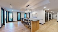 Located at 6163 Kempsville Circle in Norfolk, Virginia, this thoughtfully designed commercial suite offers a refined blend of modern aesthetics and functional layout across a single floor. Light wide-plank hardwood flooring flows throughout, anchoring the space with warmth and continuity.

The central lounge and gathering area is anchored by a striking bar-height island with a dark stone countertop and natural wood base, illuminated by a rectangular crystal chandelier set within a coffered ceiling detail. Black-framed glass doors and recessed lighting add a clean, contemporary touch, while built-in cabinetry with dark stone tops along a whitewashed exposed brick wall provides both storage and visual character. Nautical-inspired décor, including framed artwork and decorative oars, nods to the coastal Virginia setting.

The dining area is a standout feature, enclosed by floor-to-ceiling black-framed windows that invite abundant natural light and offer open views of the surrounding landscape. A rustic wood dining table is complemented by upholstered seating and a modern multi-arm chandelier, creating an inviting space ideal for meetings or collaborative gatherings. The whitewashed brick walls and wall-mounted display screen further enhance its versatility. Additional amenities include a dedicated bathroom, laundry room, and a utility alcove housing office equipment.