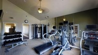 This single-floor fitness center is thoughtfully designed to provide a comprehensive workout environment. The space features vaulted ceilings that create an open, airy atmosphere, complemented by a ceiling fan and warm pendant lighting that keeps the room comfortable and well-lit during exercise sessions. A wall of floor-to-ceiling mirrors lines one side of the room, visually expanding the space and allowing users to monitor their form throughout their workouts. The cardio zone is equipped with treadmills, elliptical machines, and stair climbers, each fitted with individual display screens for entertainment and performance tracking. A multi-station cable strength training machine is positioned toward the back of the room, offering a variety of resistance-based exercises. The flooring consists of durable, low-pile carpet suitable for a fitness environment, while olive-toned accent walls add a modern, polished aesthetic. Wall-mounted televisions provide additional entertainment options. A wall clock is conveniently placed for time management during workouts. An entry door with a sidelight window connects the fitness center to the rest of the building, ensuring easy accessibility. Overall, the space strikes a balance between functionality and design, making it a well-appointed facility for a full-body fitness routine.