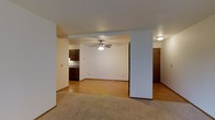 This single-floor space showcases an open, flowing layout that connects the main living area to the kitchen and additional rooms. The living area is finished with wall-to-wall carpet, providing a comfortable underfoot feel, while the adjoining space transitions to warm hardwood-style flooring that extends toward the kitchen. White walls throughout create a clean, neutral backdrop, and a ceiling fan with a built-in light fixture serves as both a functional and understated focal point in the central area.

A structural column adds architectural definition, subtly separating the living space from the adjacent area without fully closing off the open feel. The kitchen, visible in the background, features dark-toned cabinetry paired with light countertops, offering a classic contrast. A doorway nearby hints at additional rooms, including what appears to be a hallway leading to further living quarters. Flush-mount lighting provides consistent illumination throughout. The overall ambiance is clean, practical, and straightforward, offering a versatile foundation well-suited for a variety of interior arrangements and personal styles.