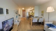 This well-designed single-floor residence showcases a thoughtfully planned open-concept layout that brings together the living, dining, and kitchen areas into one cohesive and functional space. Light hardwood-style flooring runs throughout, lending warmth and continuity to the interior.

The kitchen is outfitted with modern stainless steel appliances, including a full-size refrigerator and microwave, complemented by sleek two-tone cabinetry featuring white upper cabinets and dark wood lower cabinets. A subway tile backsplash adds a clean, contemporary touch, while a small breakfast bar with industrial-style stools offers a casual dining option.

The dining area accommodates a full rectangular wood table surrounded by upholstered chairs, making it ideal for both everyday meals and entertaining. Adjacent to it, the living area features a media console with a flat-screen television and a white open-frame shelving unit that adds a decorative yet functional element to the space.

Recessed lighting illuminates the interior throughout, and neutral wall tones contribute to the bright and airy ambiance. A wood-panel entry door anchors the far end of the space, providing a clear sense of flow from entry to living area. The overall aesthetic is modern, clean, and move-in ready.