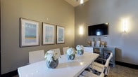 This conference room offers a polished and professional environment designed to facilitate productive meetings and collaborative gatherings. The space is defined by impressively tall ceilings that lend an open, airy quality, enhanced by a combination of overhead pendant lighting and wall-mounted sconces that cast a warm, inviting glow throughout.

At the center of the room sits a long, high-gloss white conference table surrounded by sleek white and chrome chairs, providing a clean and contemporary aesthetic. Fresh white floral arrangements in geometric vases are arranged along the table, adding an elegant, refined touch to the workspace.

The walls, finished in a warm taupe tone, serve as a sophisticated backdrop for a curated selection of framed artwork, including a triptych of abstract prints in cool blue and green hues, as well as a bold turquoise abstract piece in a gold frame. A wall-mounted flat-screen display positioned above a stylish credenza makes the space well-equipped for presentations and media sharing.

Natural light filters in through dark-trimmed windows, offering views of an outdoor area featuring stone accents and lush greenery. A tall decorative plant in the corner adds a lively, organic element to the space. Textured carpet flooring and dark baseboards complete the room's cohesive, upscale design, making it a distinguished setting for any professional occasion.