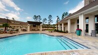 This community pool area in Panama City Beach, Florida, offers a thoughtfully designed outdoor retreat that blends functional amenities with resort-style aesthetics. The centerpiece is a generously sized, freeform swimming pool with crystal-clear blue water, accessible via wide entry steps with handrails and framed by an elegant paver deck that extends throughout the entire outdoor space.

A covered pergola pavilion anchors one side of the pool deck, equipped with a mounted television and surrounded by ample seating, making it ideal for both relaxed lounging and social gatherings. Lounge chairs and seating areas are distributed across the sun deck, complemented by lush landscaping including palm trees, flowering planters, and manicured greenery that add vibrant color and a tropical feel.

Adjacent to the pool is a well-appointed single-story clubhouse building, featuring large floor-to-ceiling windows, a covered porch with classic rocking chairs, and tile flooring. Inside, visible through the glass entry doors, is an inviting lounge space with a stone fireplace and comfortable furnishings. The exterior of the building is finished in crisp white with neutral trim, maintaining a clean, modern coastal character. The surrounding community features multi-story residential buildings set against a backdrop of tall pines, reinforcing the serene, nature-adjacent atmosphere of this Panama City Beach destination.