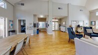 Located at 1801 Aviation Way in Redondo Beach, California, this single-story clubhouse serves as a welcoming community hub designed for both relaxation and social gathering. The space is defined by its soaring vaulted ceilings and an abundance of natural light streaming through rows of large windows and clerestory windows positioned high along the walls, creating an open and airy atmosphere throughout.

The main gathering area features warm hardwood flooring and flexible seating arrangements, including comfortable sofas and accent chairs, as well as large wooden communal tables suitable for group activities or casual dining. A set of French doors lines one wall, opening directly to an outdoor patio area and allowing seamless indoor-outdoor flow with views of palm trees and the surrounding landscaping.

A distinct lounge area is anchored by a tiled fireplace and a wall-mounted television, flanked by built-in white bookshelves filled with books and decorative accents. A classic upright piano nestled between the shelves adds a cultural touch, while teal-painted walls lend warmth and character to this corner of the space.

The entry and common corridor area offers high ceilings, mailbox stations, informational displays, and thoughtful seating arrangements, providing a functional and organized welcome. Throughout the clubhouse, the design emphasizes openness, community, and comfort, making it an ideal gathering place for residents.