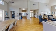 Located at 1801 Aviation Way in Redondo Beach, California, this single-story clubhouse serves as a welcoming community hub designed for both relaxation and social gathering. The space is defined by its soaring vaulted ceilings and an abundance of natural light streaming through rows of large windows and clerestory windows positioned high along the walls, creating an open and airy atmosphere throughout.

The main gathering area features warm hardwood flooring and flexible seating arrangements, including comfortable sofas and accent chairs, as well as large wooden communal tables suitable for group activities or casual dining. A set of French doors lines one wall, opening directly to an outdoor patio area and allowing seamless indoor-outdoor flow with views of palm trees and the surrounding landscaping.

A distinct lounge area is anchored by a tiled fireplace and a wall-mounted television, flanked by built-in white bookshelves filled with books and decorative accents. A classic upright piano nestled between the shelves adds a cultural touch, while teal-painted walls lend warmth and character to this corner of the space.

The entry and common corridor area offers high ceilings, mailbox stations, informational displays, and thoughtful seating arrangements, providing a functional and organized welcome. Throughout the clubhouse, the design emphasizes openness, community, and comfort, making it an ideal gathering place for residents.