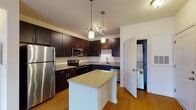 This single-floor apartment at Spring House Apartments offers a thoughtfully designed layout with modern finishes and functional living spaces. The kitchen serves as a central highlight, featuring rich dark-espresso cabinetry, granite countertops, and a full suite of stainless steel appliances including a refrigerator, range, over-the-range microwave, and dishwasher. A center island with a granite surface provides additional prep space and casual seating, while pendant lighting and warm hardwood-style flooring add a welcoming ambiance. The kitchen also provides convenient in-unit laundry access through an adjacent utility closet. The bathroom is well-sized, offering hardwood-style flooring, dark cabinetry with granite countertops, a tall storage tower, and a tiled tub-shower combination. The bedroom is bright and comfortable, with plush carpet, a ceiling fan, and large windows that invite natural light and outdoor views. A generously sized walk-in closet with wire shelving provides ample storage. Throughout the apartment, the neutral color palette, clean lines, and consistent use of quality materials create a cohesive and comfortable living environment.