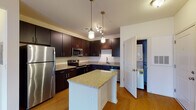 This single-floor apartment at Spring House Apartments offers a thoughtfully designed layout with modern finishes and functional living spaces. The kitchen serves as a central highlight, featuring rich dark-espresso cabinetry, granite countertops, and a full suite of stainless steel appliances including a refrigerator, range, over-the-range microwave, and dishwasher. A center island with a granite surface provides additional prep space and casual seating, while pendant lighting and warm hardwood-style flooring add a welcoming ambiance. The kitchen also provides convenient in-unit laundry access through an adjacent utility closet. The bathroom is well-sized, offering hardwood-style flooring, dark cabinetry with granite countertops, a tall storage tower, and a tiled tub-shower combination. The bedroom is bright and comfortable, with plush carpet, a ceiling fan, and large windows that invite natural light and outdoor views. A generously sized walk-in closet with wire shelving provides ample storage. Throughout the apartment, the neutral color palette, clean lines, and consistent use of quality materials create a cohesive and comfortable living environment.