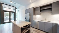 Located on Broadway in San Diego's vibrant 92101 district, this modern two-story space offers a thoughtfully designed layout spread across two floors connected by a striking steel-and-cable staircase. The ground floor features an open-plan kitchen outfitted with flat-front gray cabinetry, quartz countertops, stainless steel appliances including a French door refrigerator and dishwasher, a subway tile backsplash, and a center island with a built-in wine rack. Floor-to-ceiling glass at the entry bathes the main living area in natural light, while a ceiling fan adds comfort to the airy volume. Light wood-tone flooring flows throughout both levels, contributing to a clean, contemporary aesthetic. The upper floor offers an open living or sleeping area with large black-framed windows overlooking the streetscape, providing urban views and generous natural light. A dedicated closet with built-in shelving and hanging rods provides practical storage. Two well-appointed bathrooms feature gray vanity cabinetry, quartz countertops, large-format tile flooring, and marble-look tile surrounds. A hallway connects the upper-floor spaces efficiently. The overall design is sleek and modern, ideal for a live/work or residential configuration in the heart of San Diego.
